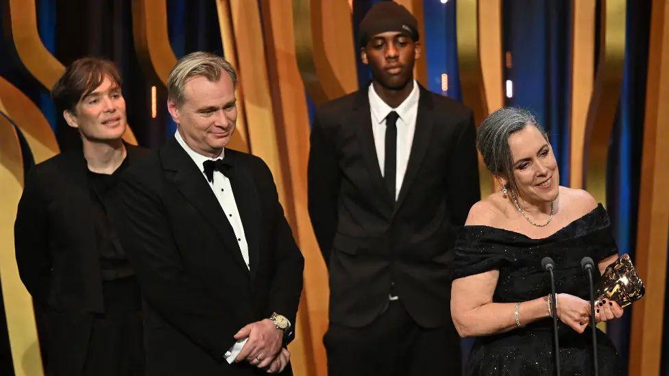 Cillian Murphy, Christopher Nolan, and Emma Thomas accept the best film award for Oppenheimer on stage during the Bafta Film Awards 2024 at the Royal Festival Hall on February 18 in London, while a YouTube prankster stands in the background