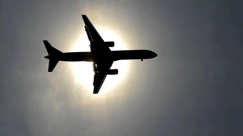 A shadowy outline of an aeroplane flies the sun, with the sky around it looking dark by comparison