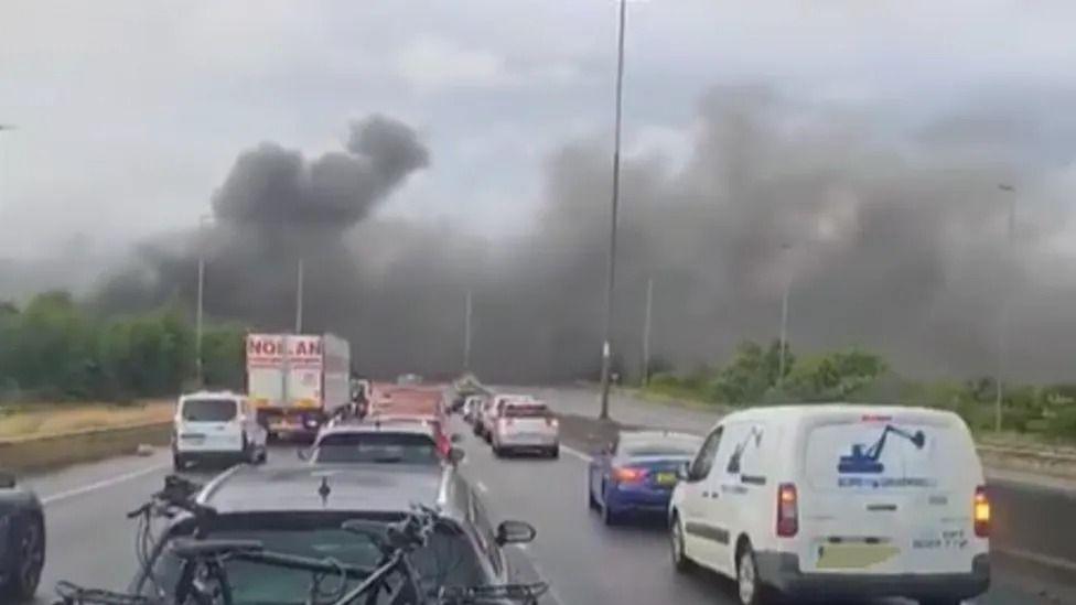 A blurry photo of a busy motorway with thick black smoke billowing across both carriageways in the distance