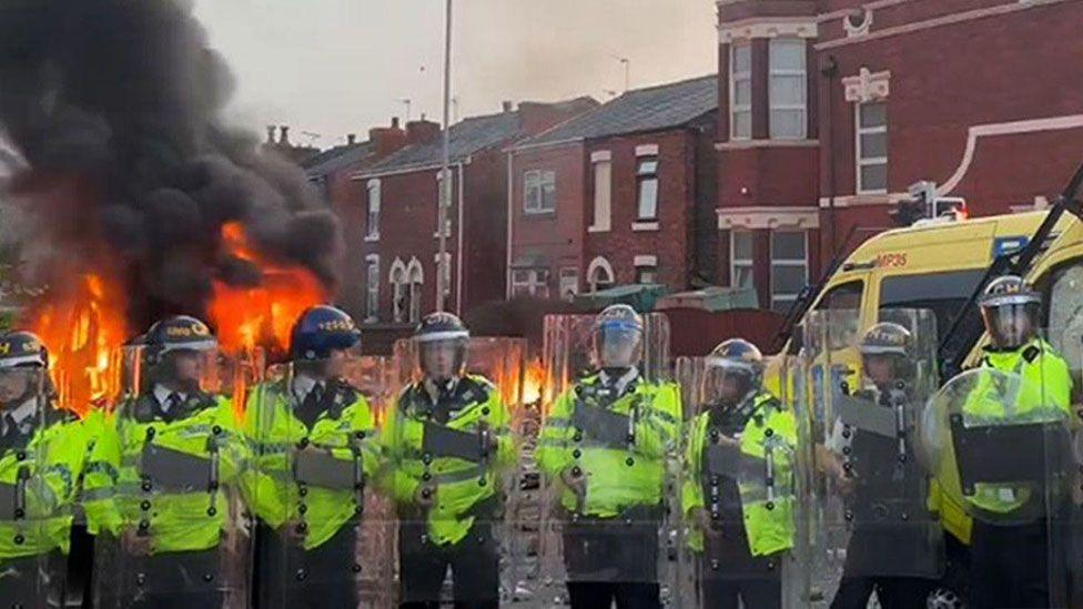 Line of riot police in high vis clothing holding riot shields outside mosque where one police van is on fire