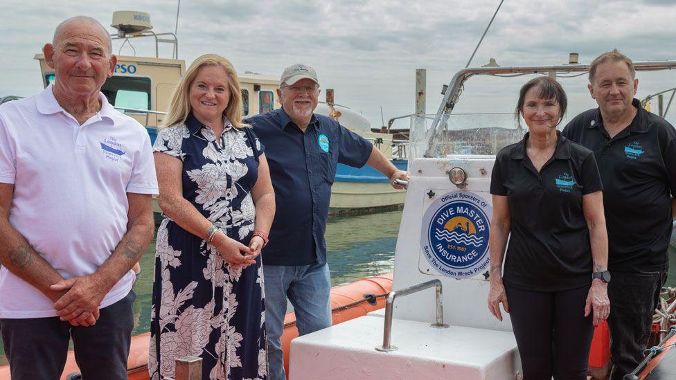 Five people, all smiling, on a boat deck. On the left is a man in a white polo shirt with a blue logo saying London Shipwreck Project, to his left is Helen Gray, from DP World, who has long blonde hair and is wearing a navy dress with large white flowers. To her left is Robert Archell, from Dive Masters Insurance, wearing a navy blue short-sleeved shirt and white cap and he has a white goatee beard. To his left is a woman with black hair pulled back from her face, wearing a black trousers and a polo-shirt with a turquoise logo saying London Shipwreck Project. To her left is Steve Ellis also dressed in a black polo-shirt with a turquoise logo on his left chest saying the London Shipwreck Project.