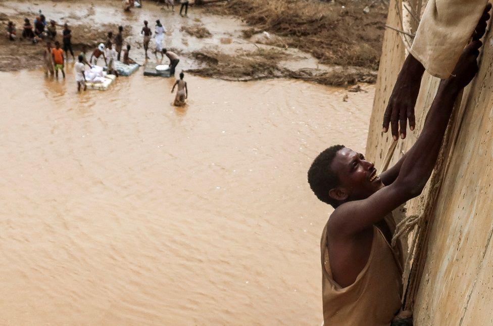 A survivor is being helped to climb a wall. Behind him is muddy, brown water.