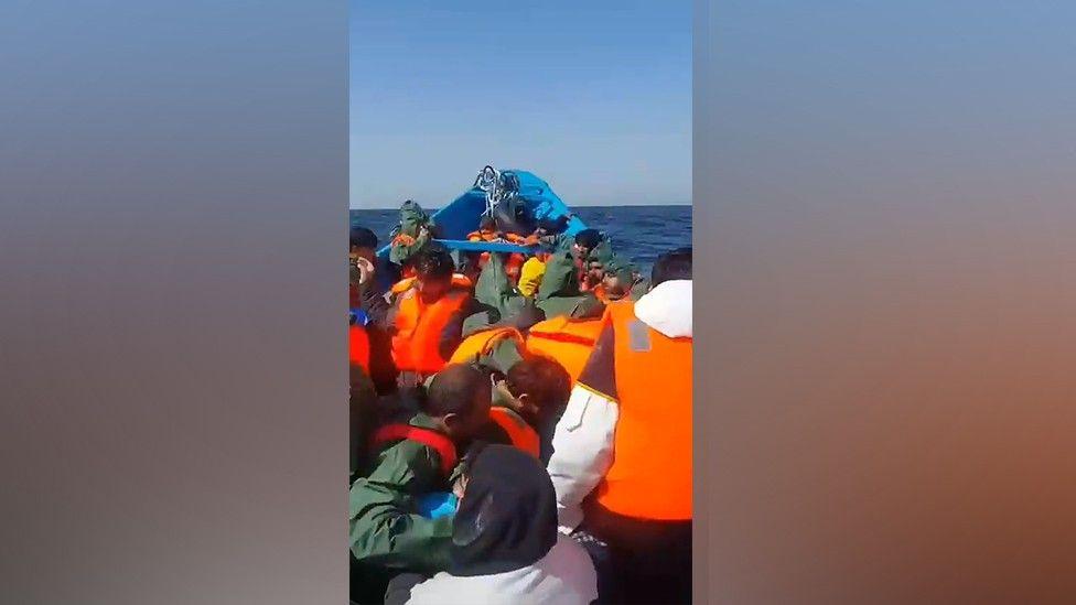 A screengrab from the boat shortly after it left Mauritania. There are many migrants wearing orange lifejackets, all facing away from the camera.