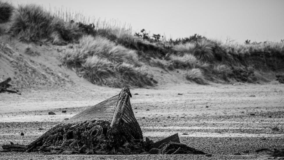 A black and white image of a beach with a wrecked old boat protruding from the sand, and dunes in the background.