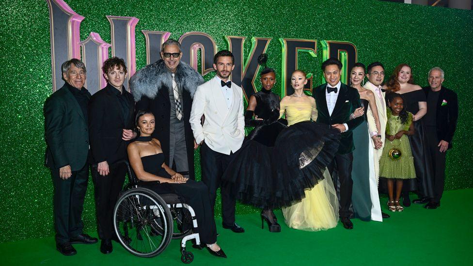 (L-R) Ethan Slater, Marissa Bode, Jeff Goldblum, Jonathan Bailey, Cynthia Erivo, Ariana Grande, Michelle Yeoh, Bowen Yang, Karis Musongole and Bronwyn James, all in smart evening wear, stand on a green carpet and in front of a green background carrying the word Wicked