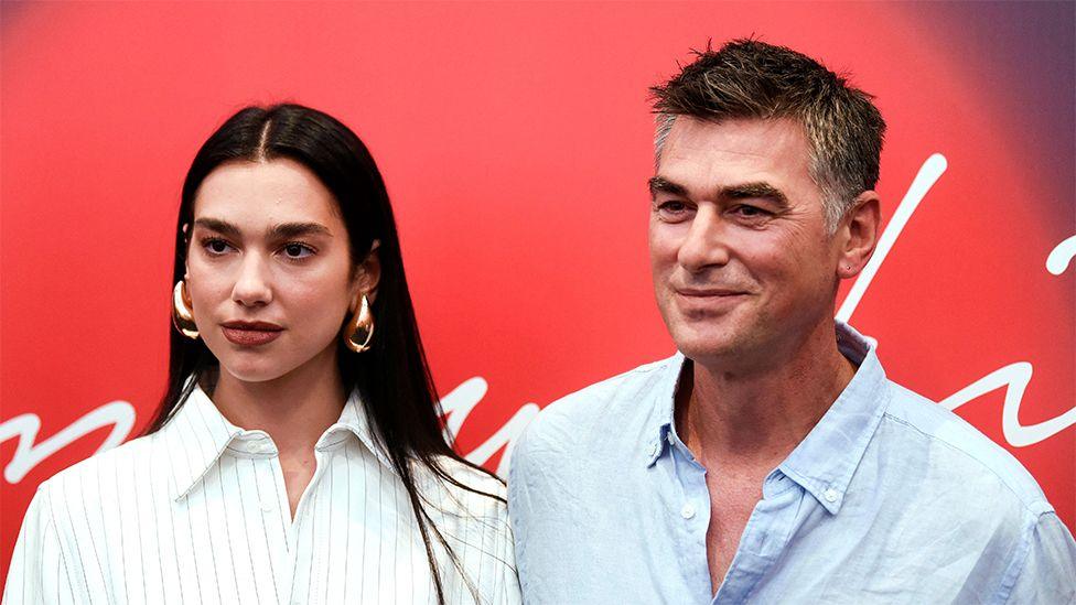 Dua Lipa, wearing a white striped shirt and her dad Dukagjin wearing a blue shirt. The background is red with white branding.