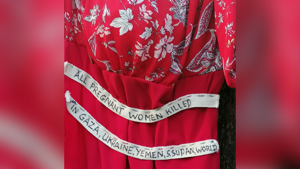 A red dress embroidered with a sash reading "All pregnant women killed in Gaza, Ukraine, Yemen, S. Sudan, World" 