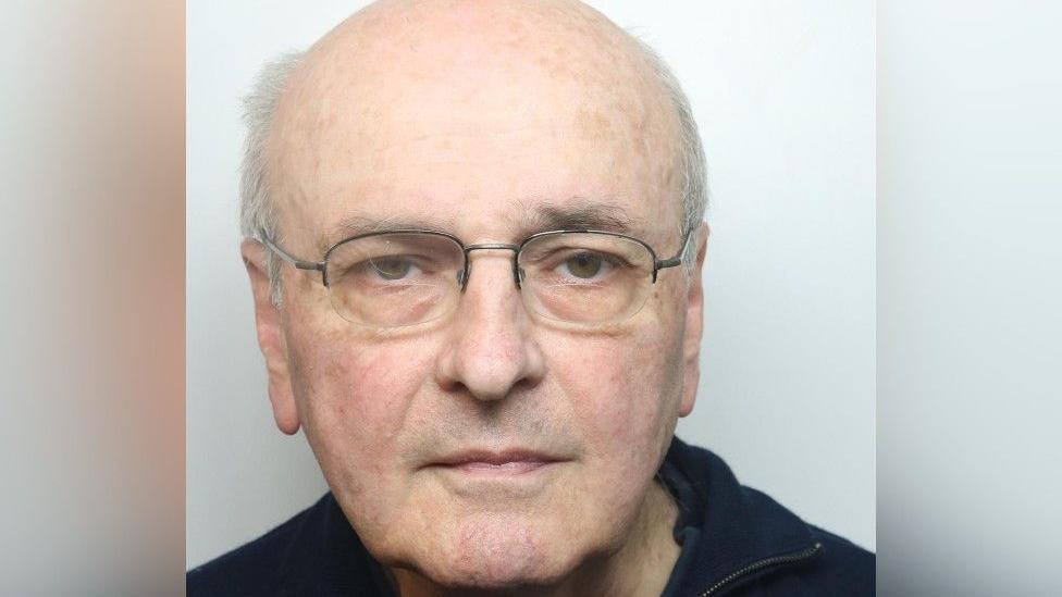 A mugshot of a man with white hair, a balding head and brown eyes. He is wearing spectacles and a navy jumper.