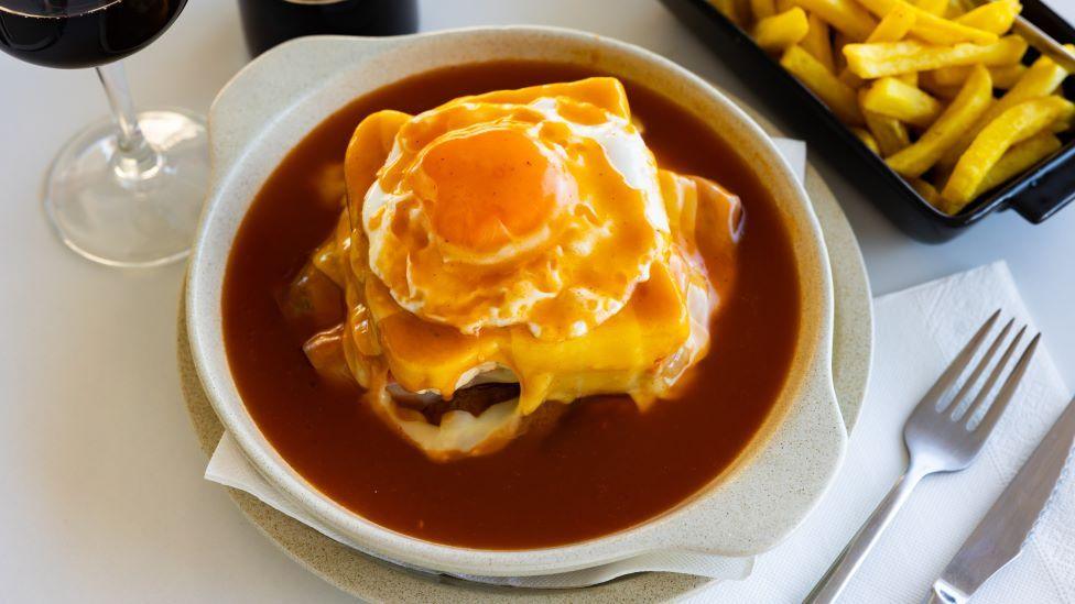 A Francesinha - a sandwich with a fried egg on top that is swimming in a gravy-like sauce - is served on a white plate on a white table, next to a tray of chips