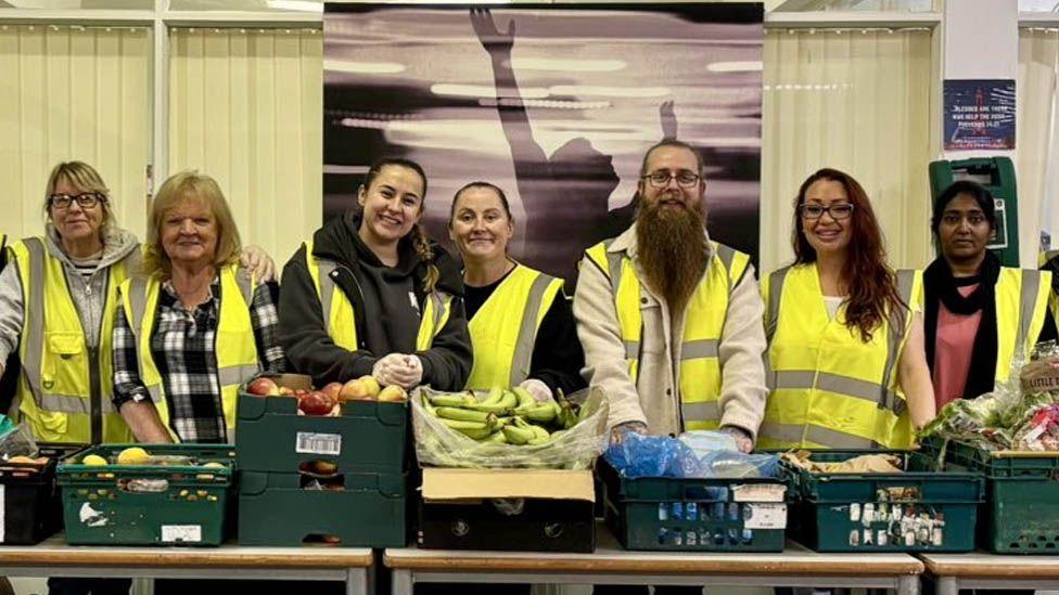Some of the volunteers at Helping Hearts, all wearing yellow hi-vis vests, stand behind a row of tables containing green plastic baskets full of food