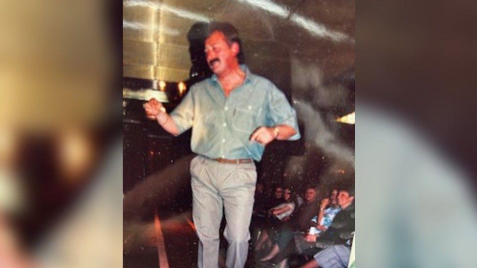 A man in a light blue shirt and light grey trousers walks down a catwalk as people watch on from the side. 