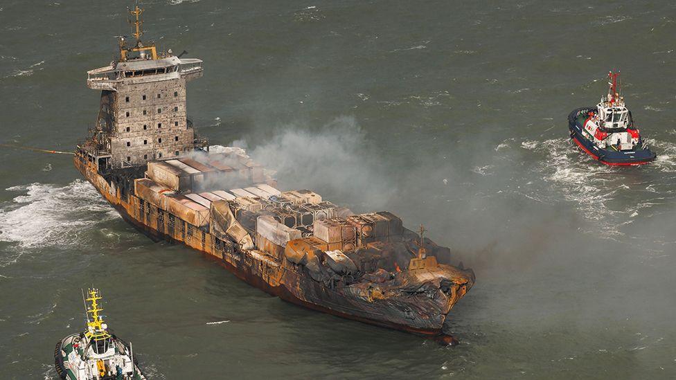 Aerial view of the Solong cargo ship with a damaged and burnt bow and surrounded by smoke with two tug boats nearby.