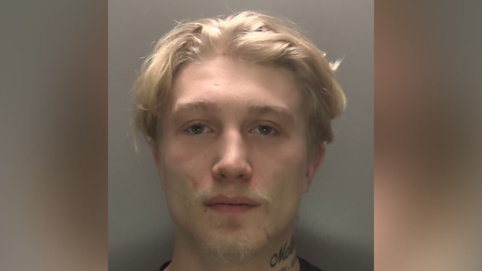 A mugshot of Cory Heritage, who has mid-length blond hair. He has a tattoo on his neck