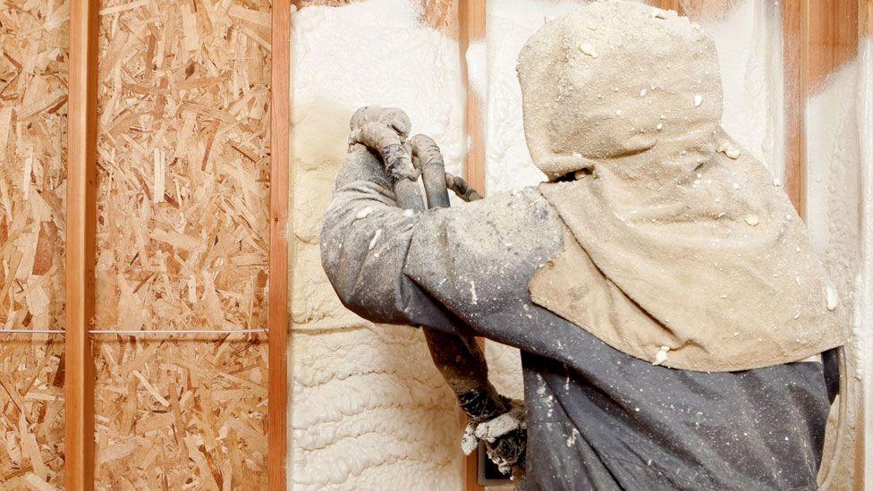 A technician in a protective blue and cream suit uses a spray gun to spray foam against a wall