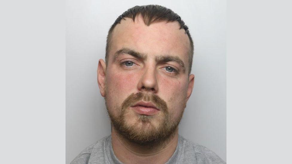 A police mugshot of Reece McKinven. He is wearing a grey t-shirt and standing against a grey background. He has blue eyes, a dark blonde beard and light brown hair which is styled forwards and shaven short on the sides.