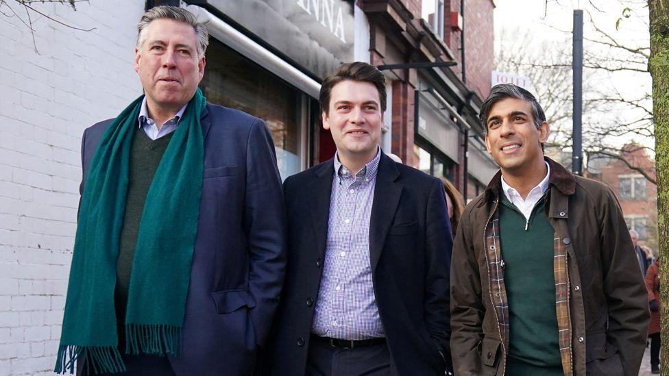 Lord Graham Brady, in a blue coat, green jumper and green scarf, and Rishi Sunak, wearing a green wax jacket, green jumper and white shirt, walk down a street on either side of a man wearing a dark blue jacket and a blue and white patterned shirt