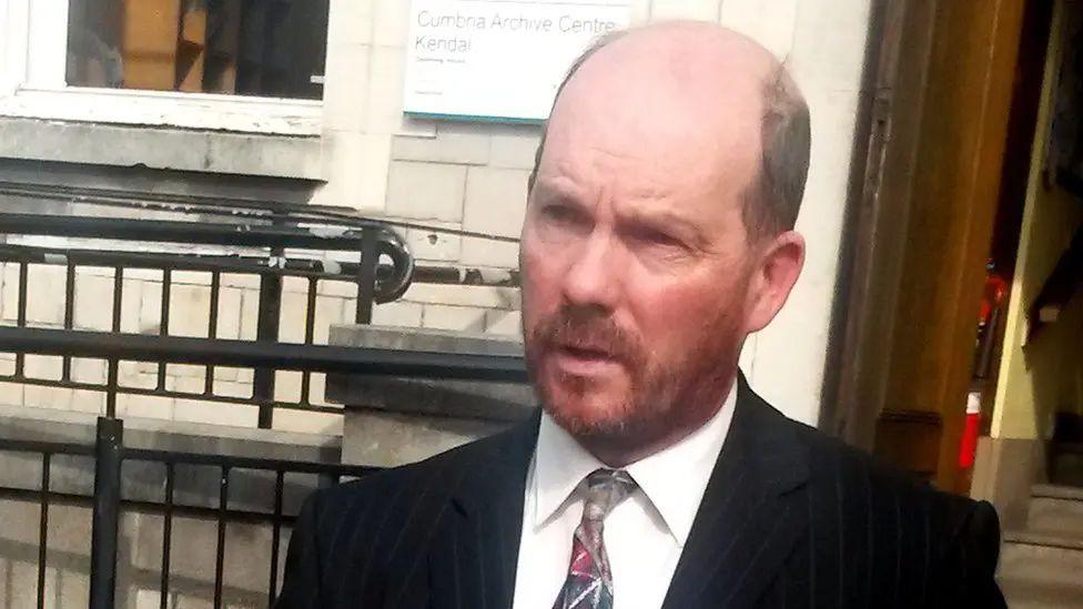 David Gill is standing outside the entrance to a building. He is wearing a suit and tie and has a bald head with dark hair at the sides and a short beard. He appears to be talking with someone off camera.