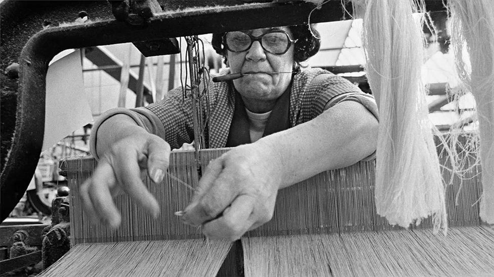 Black and white image of a weaver working at a loom in 1976. She is leaning over the loom with a tool in her mouth and she examines the threads