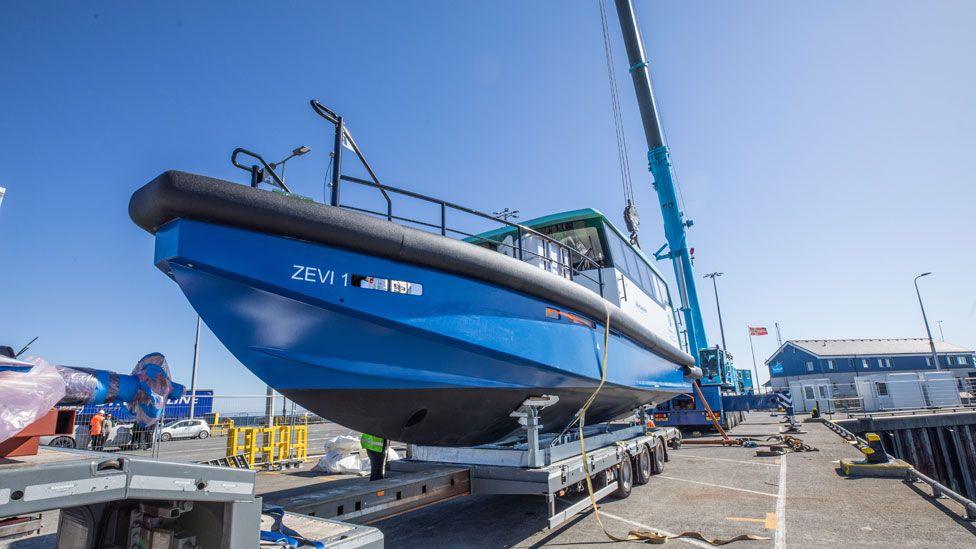 A bright blue electric boat called Zevi 1 sits on top of a wheeled metal trolley on dry land