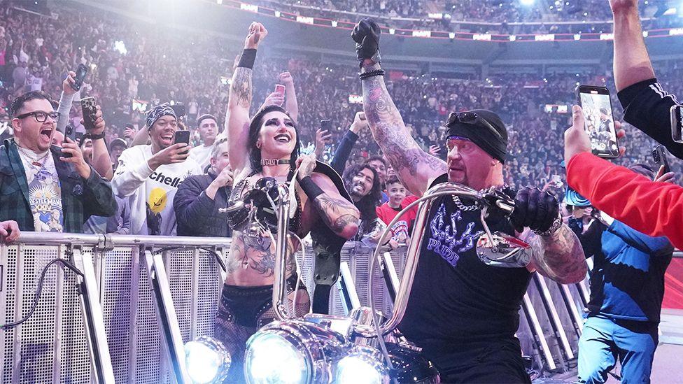 Rhea Ripley and The Undertaker, who is on a motorbike with beaming headlights. They both have their right arms raised, with fans around them taking photos and copying the pose.