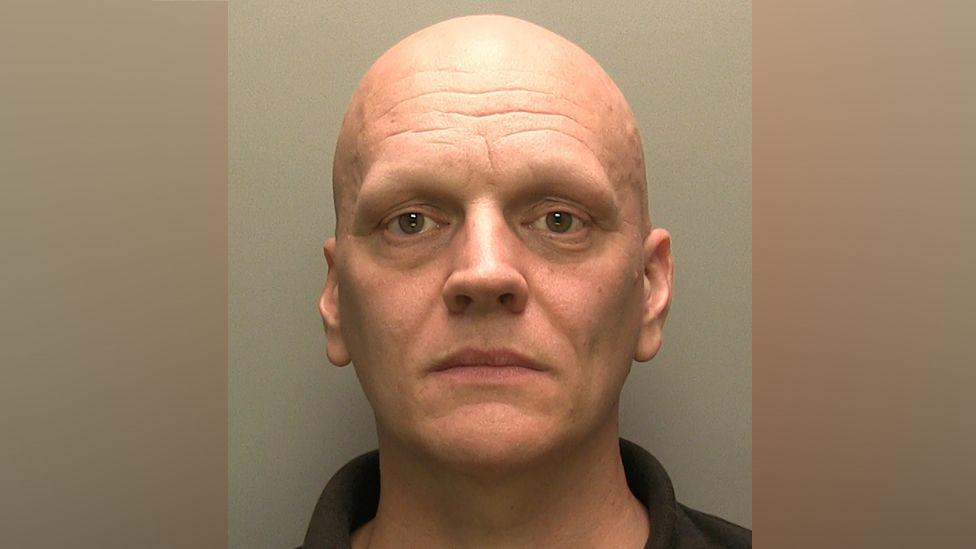 Ben Cairns has brown eyes and is clean shaven with no hair on his head.