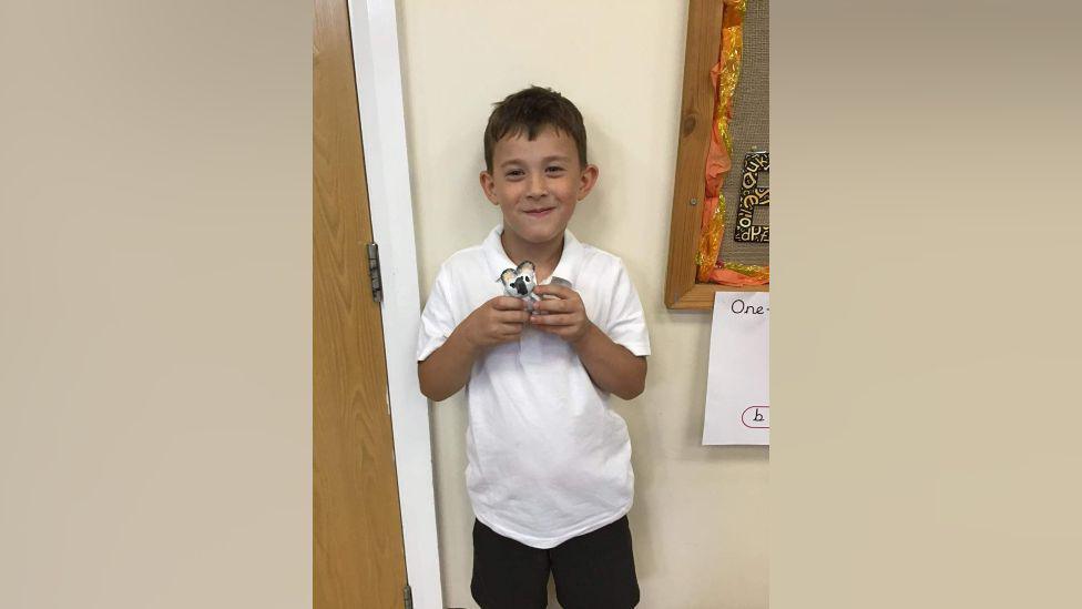 Jay Cartmell in white school T-shirt and black shorts smiling while holding a toy animal