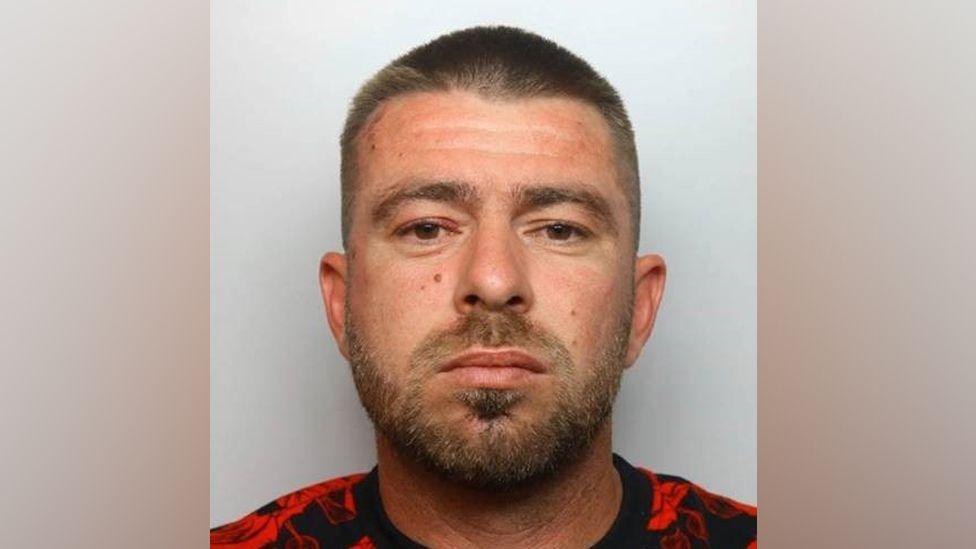 Marian Stancu mugshot against a grey background. He is white with brown eyes and has short cropped brown hair, a moustache and short beard. He is wearing a black and red t shirt.