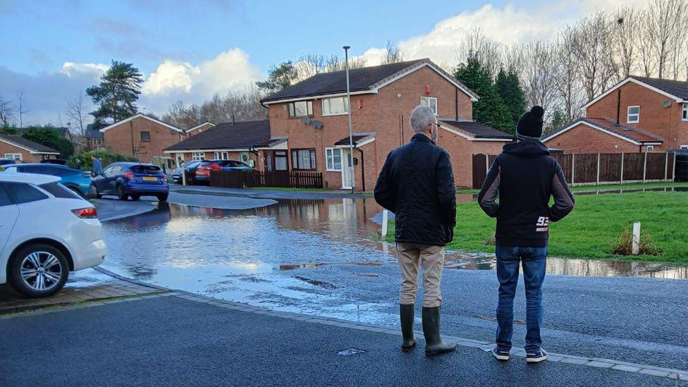 A view from behind two men stood in residential road who are facing a flooded area. One is wearing a dark jacket, beige trousers and wellies. The other is wearing a hooded top, wooly hat, blue jeans and trainers.