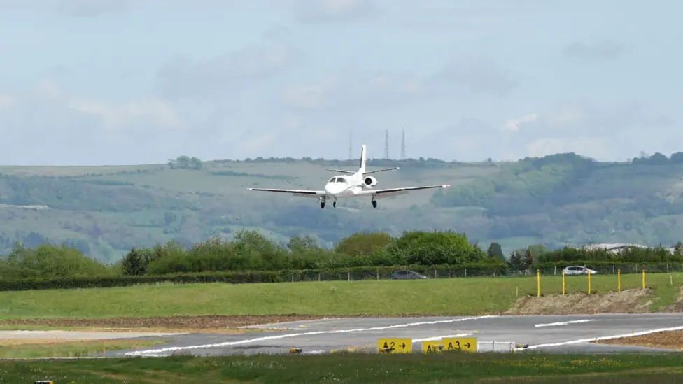 Small white plane landing on a runway at Gloucestershire Airport surrounded by grass and hills.