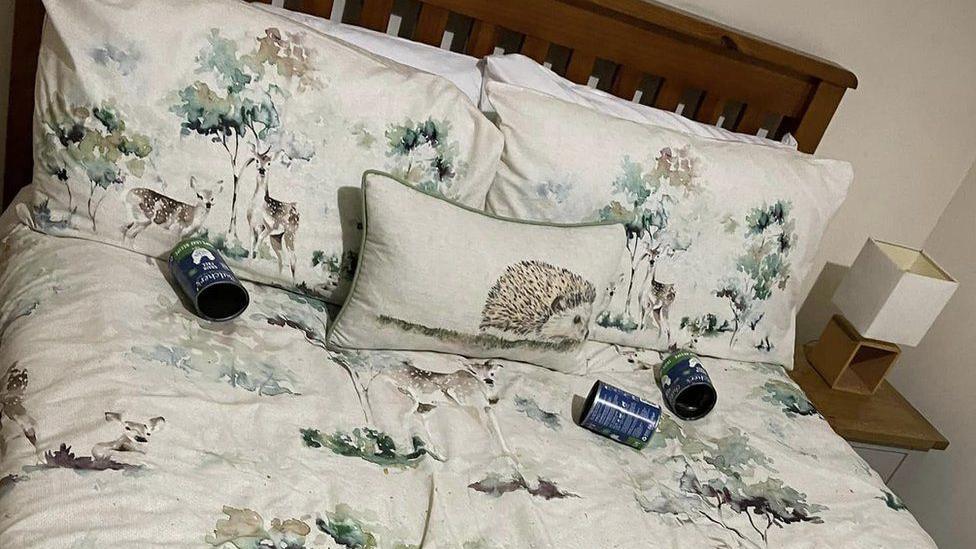 A bed with wooden frame and nice, pale, bedclothes with prints of deer and hedgehogs. There are three empty dog food cans lying on the bed.