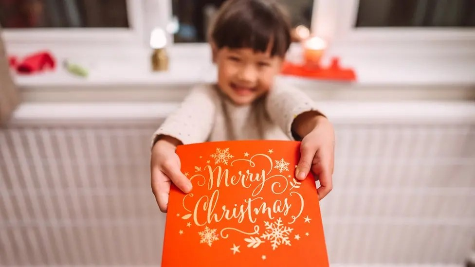 A young girl hands over a red and gold Christmas card 