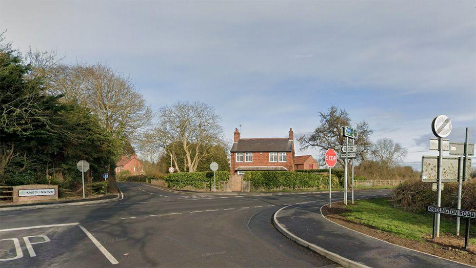 Knedlington crossroads near Howden. A red brick house stands on the corner around it are hedges and trees and road signs. A white sign for the village of Knedlington is mounted on a wall