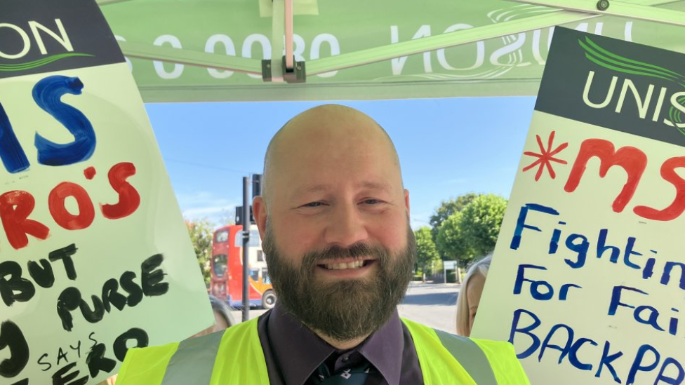 Union representative Ashley Harper, who is wearing a yellow hi-vis jacket and has a shaven head and full beard, stands on the picket line with placards either side of him. The placards have hand-written slogans such as "fighting for fair back pay".