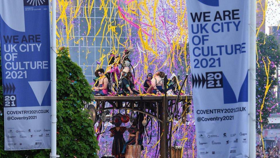 A group of dancer performing on a wooden platform in the centre of Coventry with colourful confetti and streamers in the area