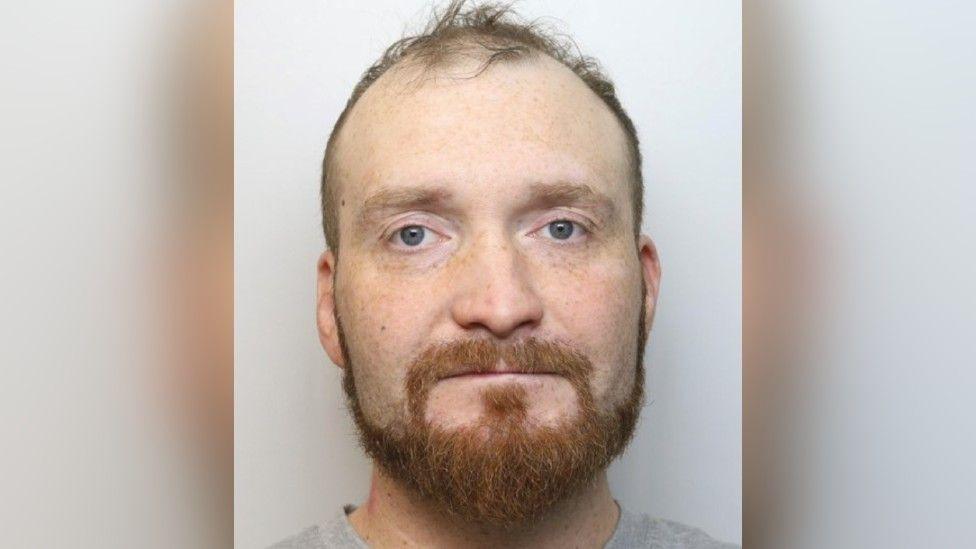 Mugshot of Marcin Janikowski. He is on a grey background and looking at hte camera with an orange moustache and beard. He has receding hairline.