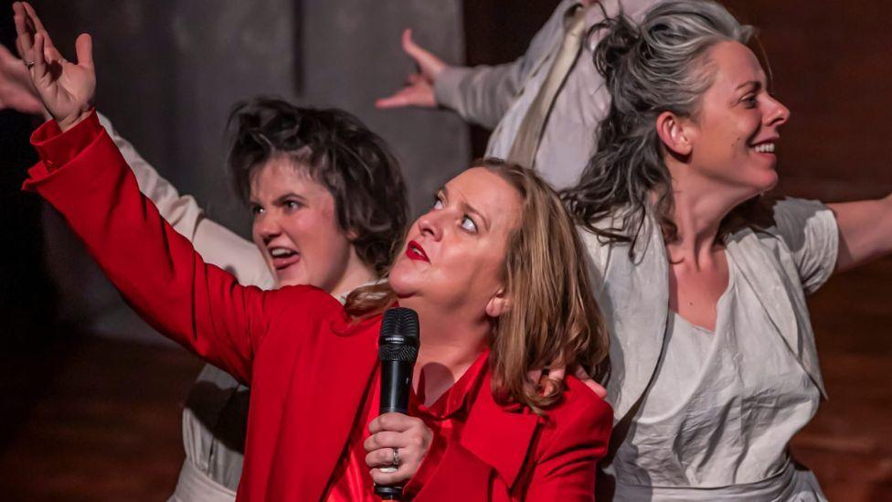 Still photography from Faust-ish play - a woman with blonde hair, in red silk pyjamas, sings into a microphone. Beside her two women, both in grey bedwear, stand close to her, looking shocked. The staging is minimal, with several sheets draped across a red brick background.