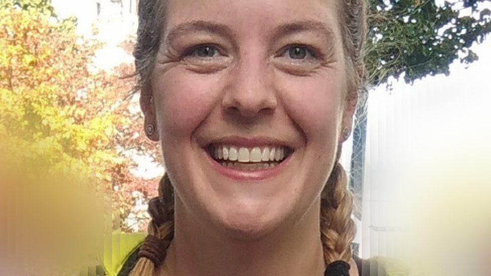 Andrea smiling at the camera, with her blond hair in two plaits. The background is blurred but trees can be seen behind her.