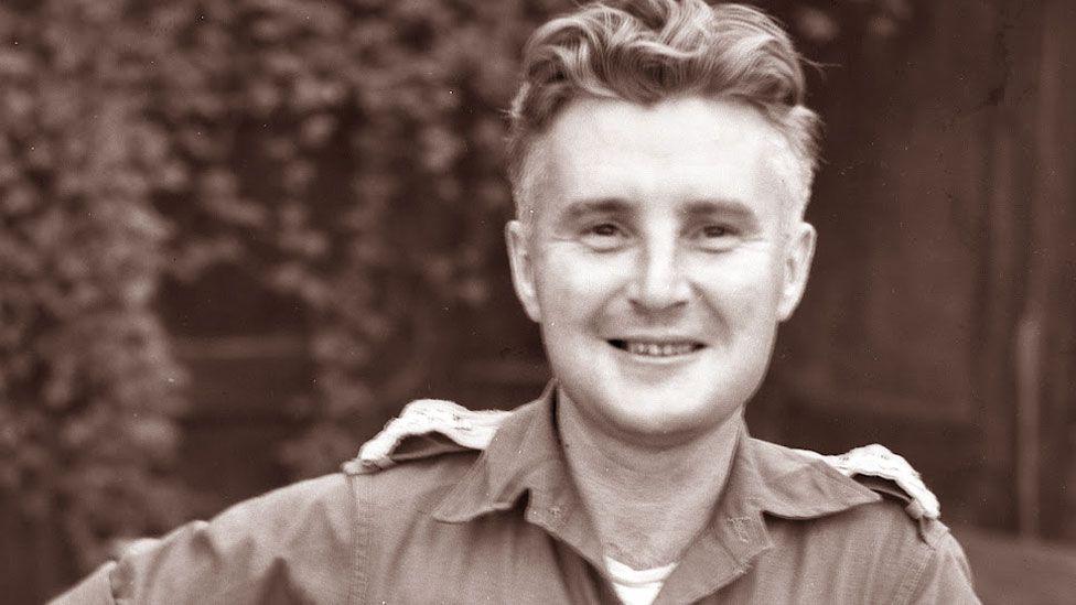 A black and white image showing Noel Duckworth in late 1945. He has fair hair, short at the sides and curling at the top and is wearing an Army uniform. He is smiling towards the camera, leaning to one side. Behind him are trailing vines and a fence. 
