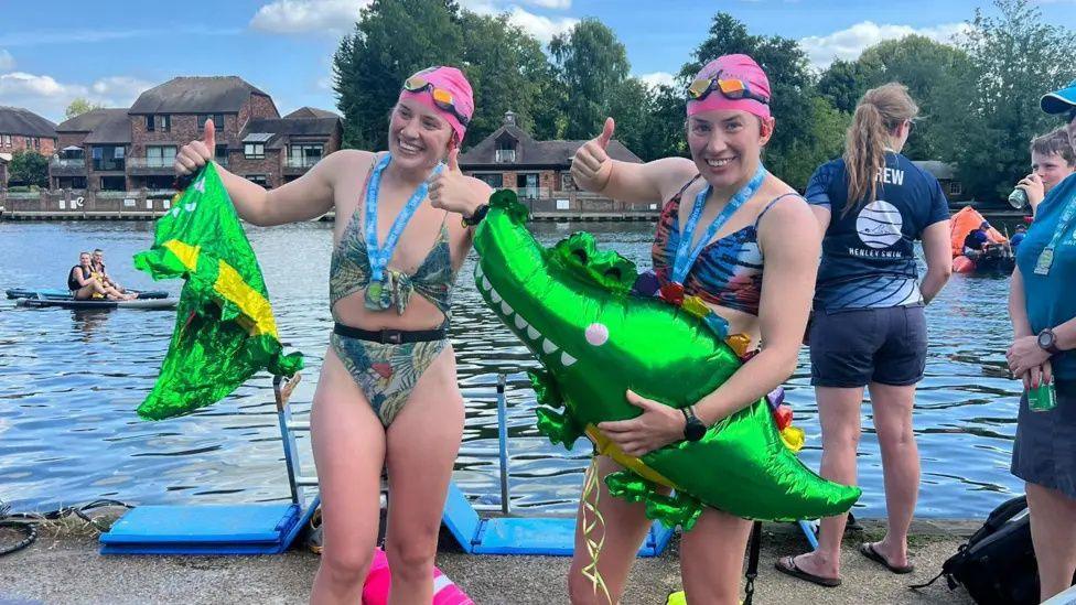 Melissa and Georgia Laurie after their swim in the Thames. They are stood in swimwear and pink caps holding inflatable green crocodile balloons.