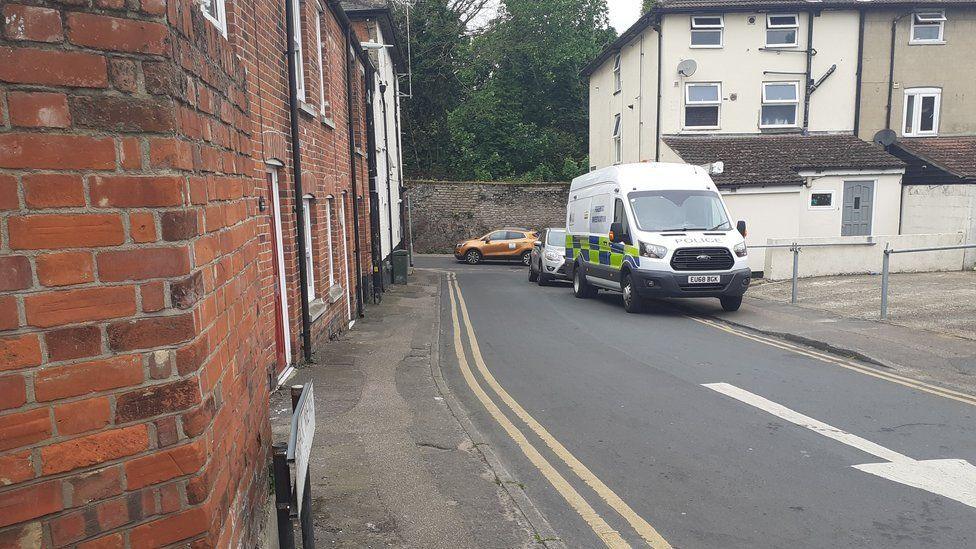 A police van parked on the pavement in a narrow residential street. There are houses either side of the road. In the background is a brick wall with trees above it.