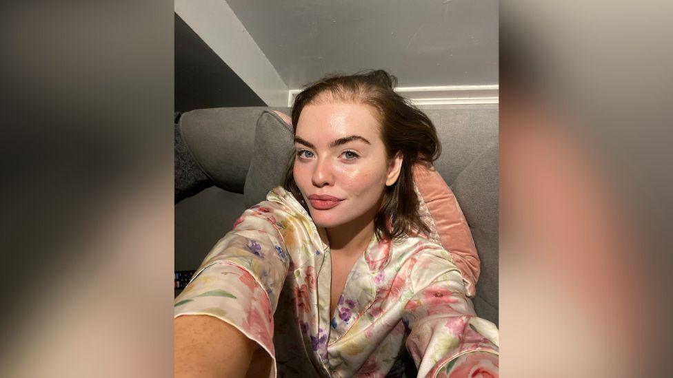 Olivia McVeigh taking a selfie. She is wearing a floral dressing gown and is sitting on a sofa. 