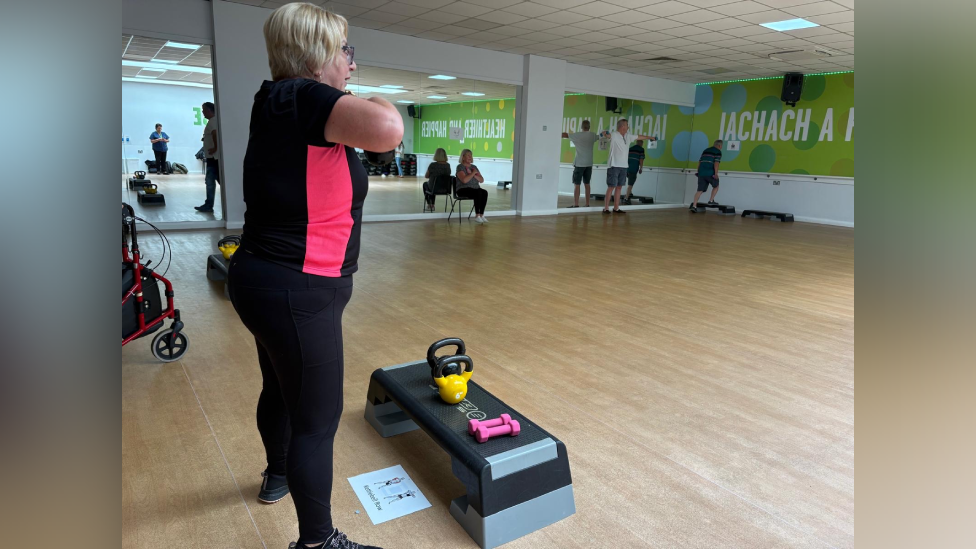 A woman with short blonde hair in black and pink gym attire, her arms are up and she is leading a fitness class. There are weights and a stepper in front of her. There are other people taking part in the class around the room. 