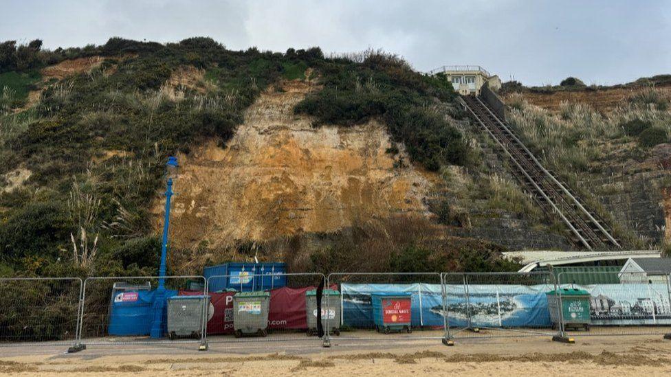 Sandy edge of beach with metal fencing, line of bins and shipping container with bare cliff seen after fall.