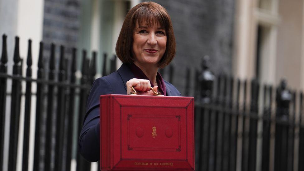 Rachel Reeves, with brown hair cut in a bob, holds a red Treasury briefcase outside Downing Street. She is smiling, standing side on to the camera and holding the briefcase in front of her body. There is a black fence in the background in front of grey-brick buildings. 