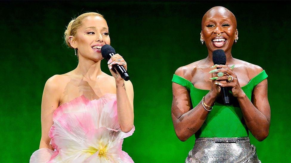 Ariana Grande and Cynthia Erivo speaking on stage. The backdrop is green. Ariana is on the left, wearing a floral dress, speaking into a black microphone. Cynthia is wearing a green top and silver shiny skirt, smiling and speaking into a black microphone.