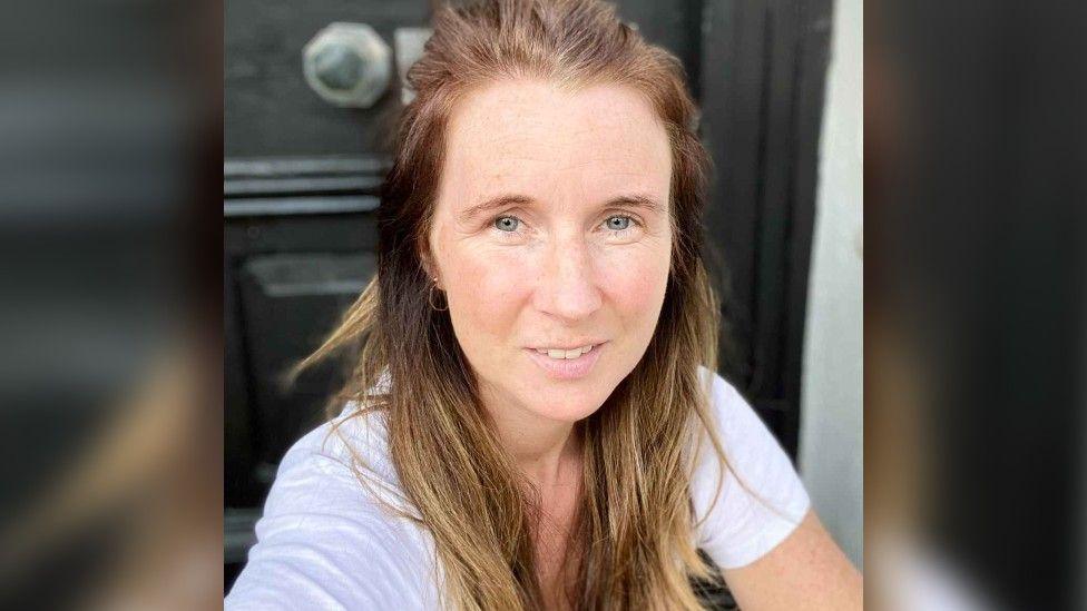 Jane Creswell is smiling at the camera, has ginger hair, blue eyes and is wearing a white t-shirt.