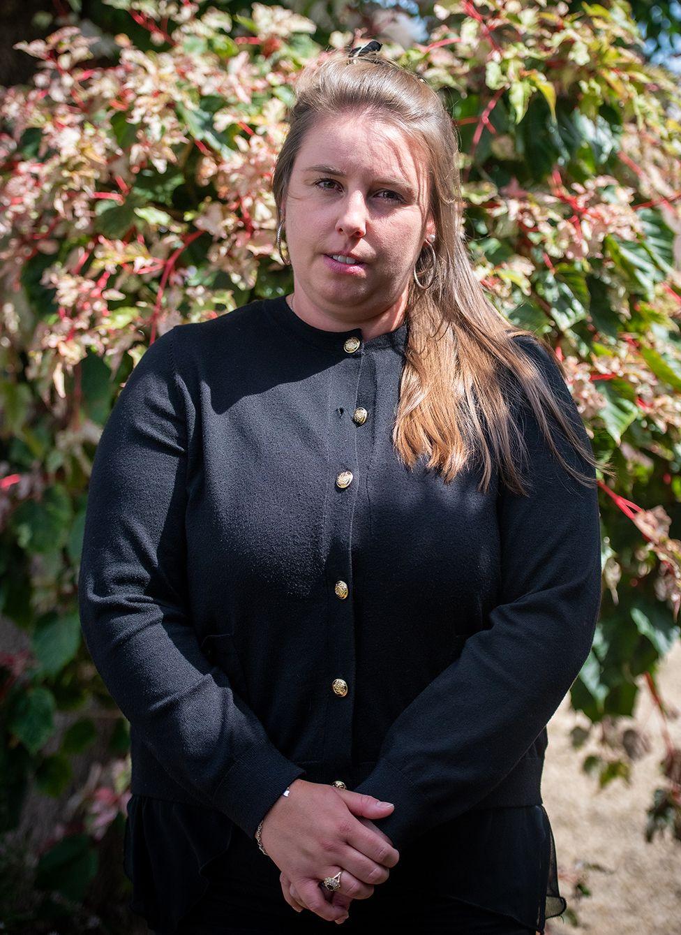 Photo of April Palmer, sister of Alice Webb who died following a Brazilian butt lift procedure one year ago. April is wearing all black clothes and is standing in the sunshine in a garden