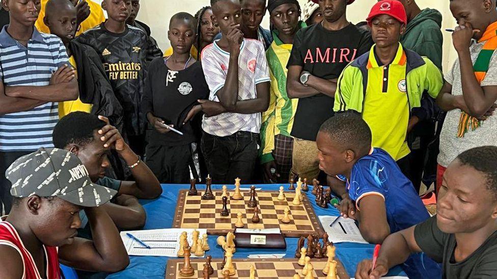 Four boys at two chess boards compete in a tournament with a crowd of young people looking on.