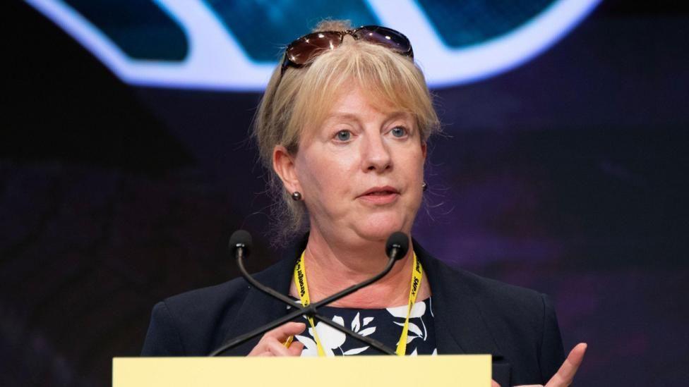 Shona Robison talks at a yellow podium with sunglasses propped on her head