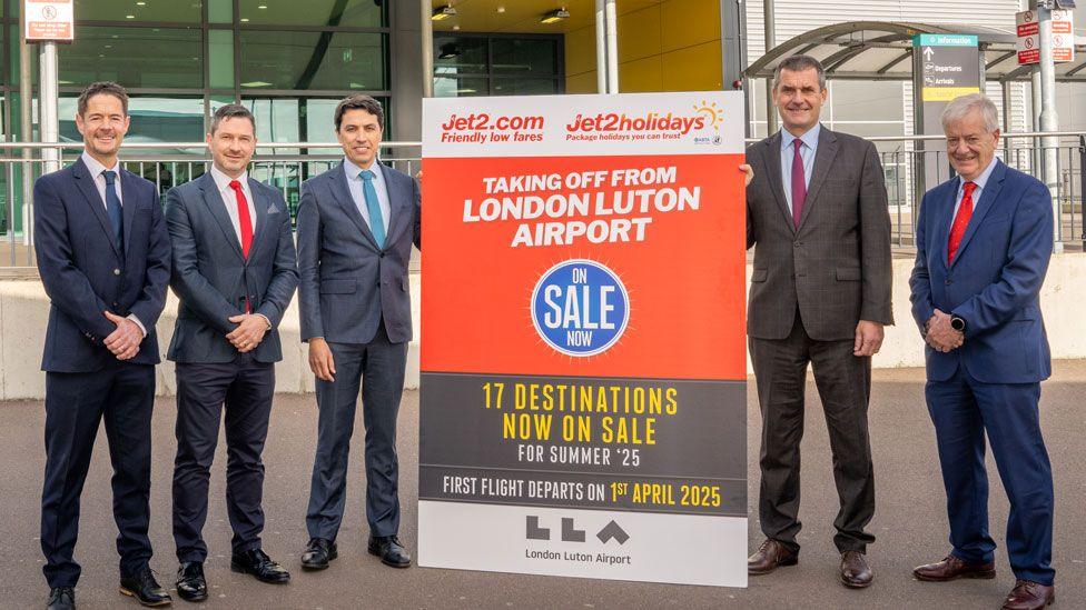 Five men in suits looking towards the camera. Between them is a sign for Jet2.com, saying it is taking off from London Luton Airport in white lettering on an orange background. They are standing outside the airport, an entry to which can be glimpsed behind them